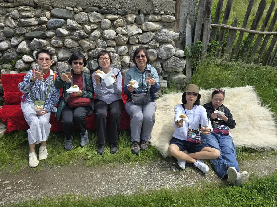 Travellers laughing at a picnic on red cushions and sheepskins, with a village lunch in hand